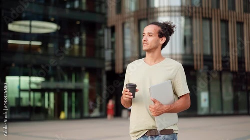 Panning medium shot of aspiring young man with laptop computer and takeaway coffee standing in city street and thinking or day dreaming of business goals achievement