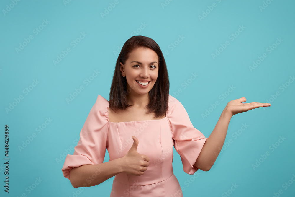 Happy broadly smiling caucasian woman with dark hair in pink dress in good mood keeps palm raised up, shows thumb up, demonstrates an attractive offer, recommends service, isolated on blue background. Happy broadly smiling caucasian woman with dark hair in pink dress in good mood keeps palm raised up, shows thumb up, demonstrates an attractive offer, recommends service, isolated on blue background.