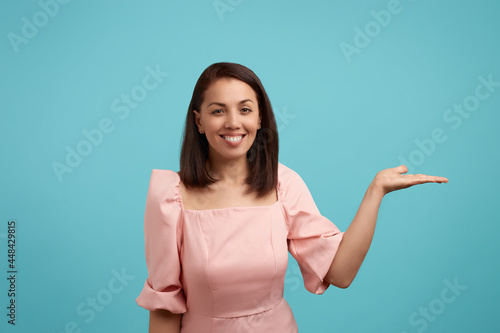 Foto Positive broadly smiling european woman with dark hair in pink dress keeps palm raised up, demonstrates result of her diligent hard work, being in high spirit, isolated over blue background