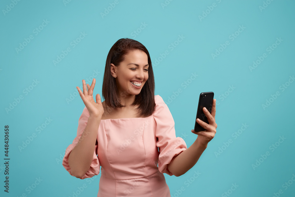 Excited european woman with dark hair and gummy smile in pink dress is laughing looking at screen of mobile phone, gestures emotionally chatting with friend, raises hand, isolated on blue background. Excited european woman with dark hair and gummy smile in pink dress is laughing looking at screen of mobile phone, gestures emotionally chatting with friend, raises hand, isolated on blue background.