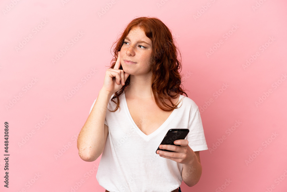 Teenager reddish woman isolated on pink background using mobile phone and thinking Teenager reddish woman isolated on pink background using mobile phone and thinking