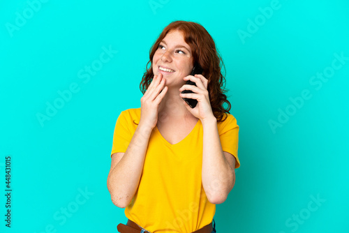 Teenager reddish woman using mobile phone isolated on blue background looking up while smiling