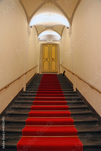 Black stone staircase, with red carpet in the center, leading to a closed decoreted door in an ancient building. Rolli palace, Genoa, Italy