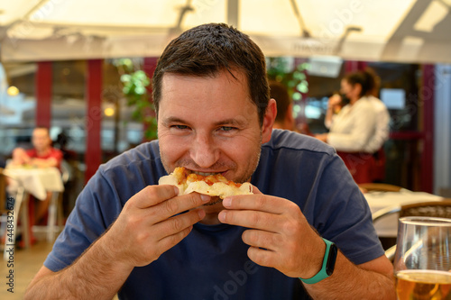 Mature man eating a slice of pizza in a restaurant. Medium shot of smiling man looking at the camera