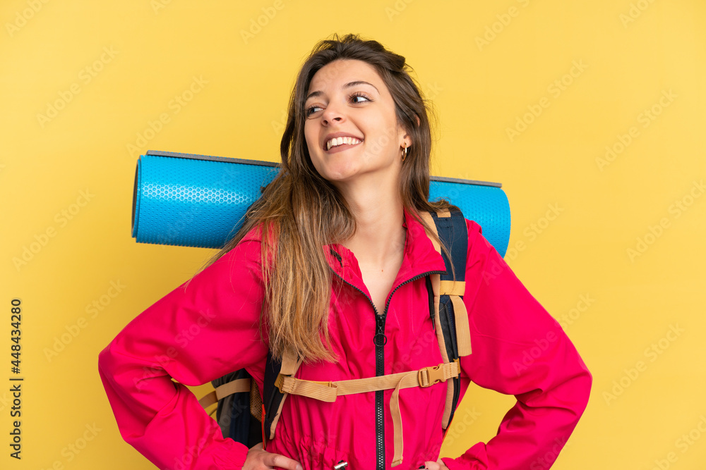 Young mountaineer man with a big backpack isolated on yellow background posing with arms at hip and smiling Young mountaineer man with a big backpack isolated on yellow background posing with arms at hip and smiling