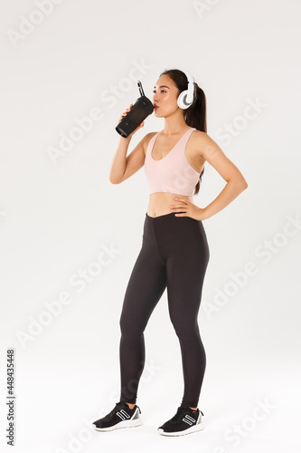 Full length of sweaty pleased female athelte drinking water from bottle while running, prepare to marathon. Fitness girl in sportswear, listening music headphones during workout, white background photo