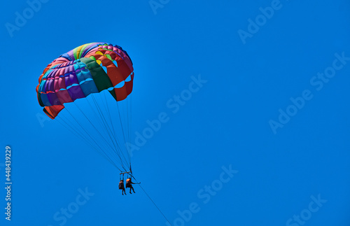 Two people, tourists flying onrainbow-colored parachute in blue sky.