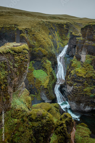 Epic aerial view of Fjaðrárgljúfur Canyon waterfall in southern Iceland. A green lush canyon with steep rocks carved into picturesque landscape in moody hazy atmosphere.