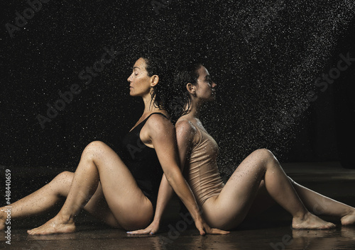 two beautiful women of Caucasian appearance with black hair sit back to back in drops of water on a black background