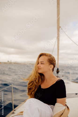 A beautiful long-haired girl sits on the bow of a barred yacht and enjoys life. The blonde is resting in the open sea or ocean.
