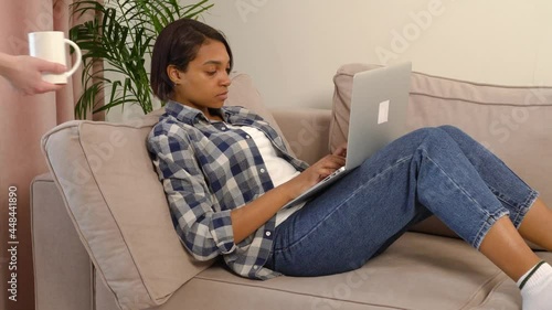 A young African-American woman works at home online using a laptop, a hand hands her a cup of coffee. Home delivery in hand. The concept of helping and delivering food home during a pandemic.