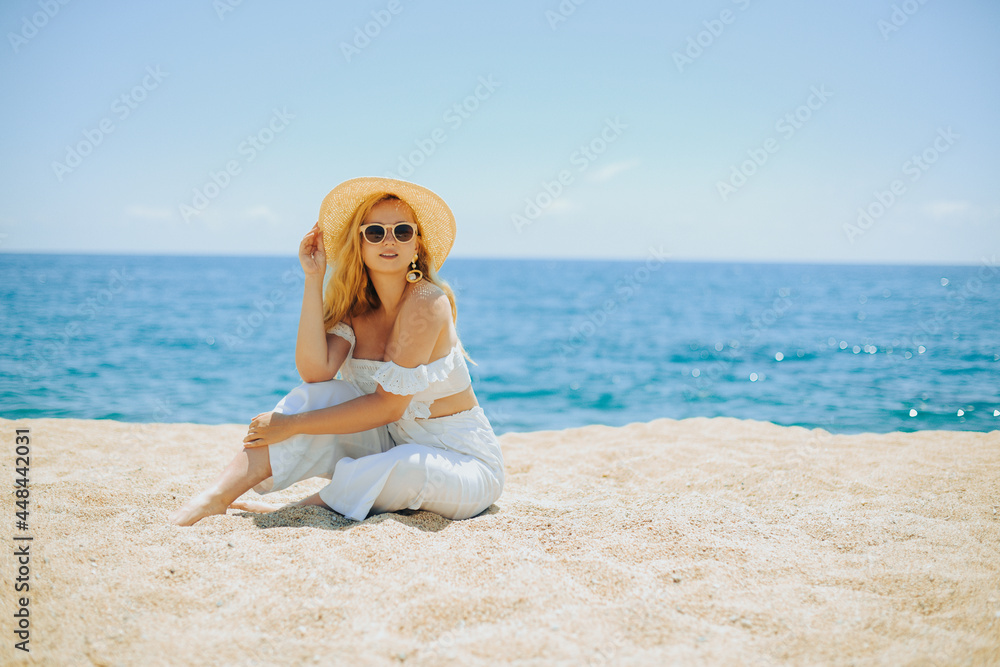 A chic fashionable long-legged model in a white outfit sits on the beach against the backdrop of the ocean and enjoys life. Vacation, vacation, summer mood. Blonde in beautiful jewelry, rich woman A chic fashionable long-legged model in a white outfit sits on the beach against the backdrop of the ocean and enjoys life. Vacation, vacation, summer mood. Blonde in beautiful jewelry, rich woman