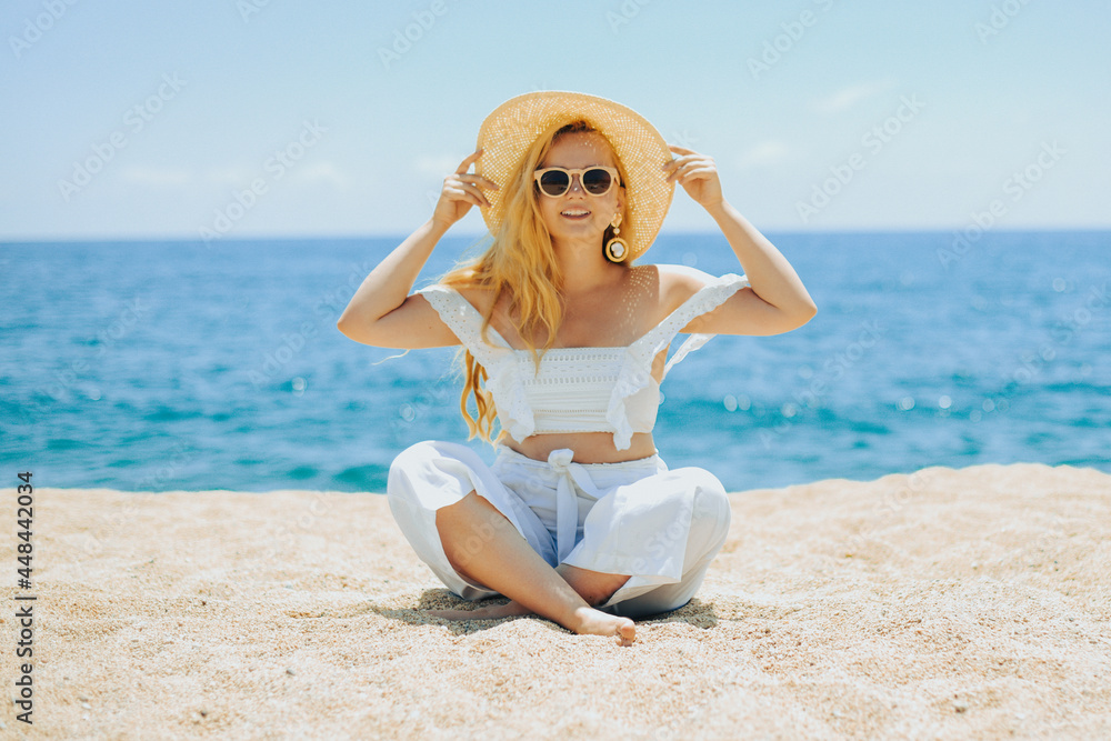 Happy girl in white outfit sits on sand against the backdrop of the sea or ocean beach. Woman smiles and laughs, vacation and joy. Fashion model, beautiful jewelry, earrings, hat, sun glasses