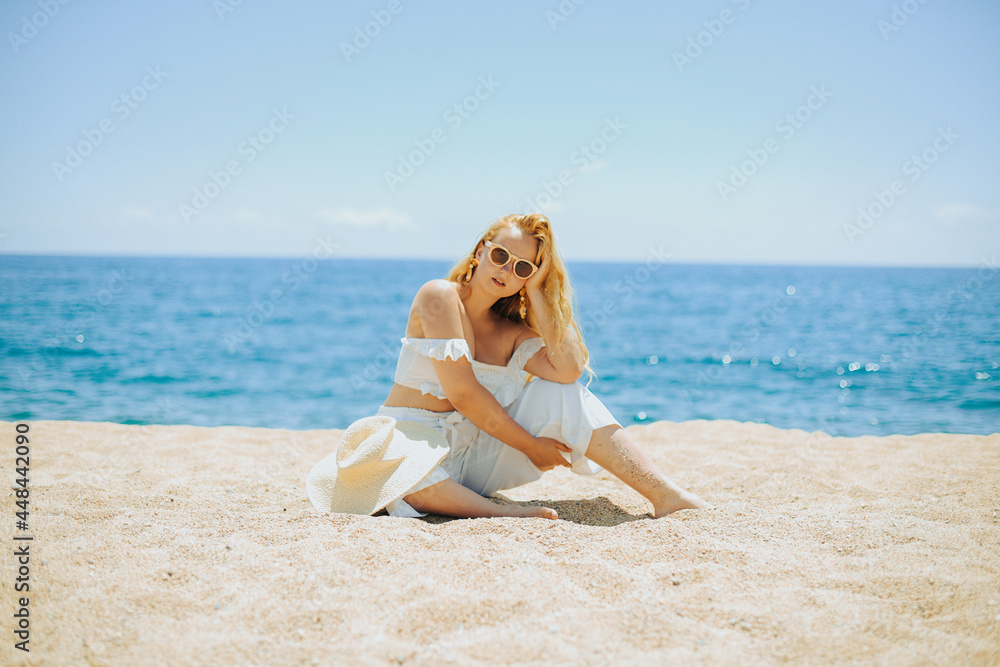 A chic fashionable long-legged model in a white outfit sits on the beach against the backdrop of the ocean and enjoys life. Vacation, vacation, summer mood. Blonde in beautiful jewelry, rich woman A chic fashionable long-legged model in a white outfit sits on the beach against the backdrop of the ocean and enjoys life. Vacation, vacation, summer mood. Blonde in beautiful jewelry, rich woman