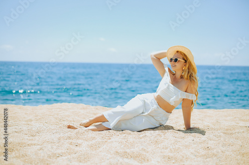 Happy girl in white outfit sits on sand against the backdrop of the sea or ocean beach. Woman smiles and laughs, vacation and joy. Fashion model, beautiful jewelry, earrings, hat, sun glasses