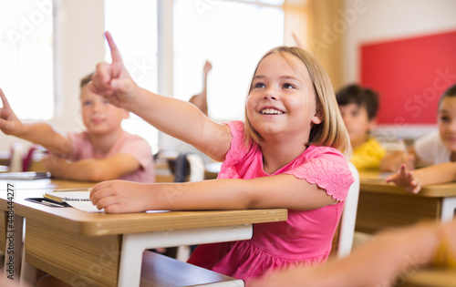 Portrait of positive pupil girl raising hand to answer during lesson at classroom