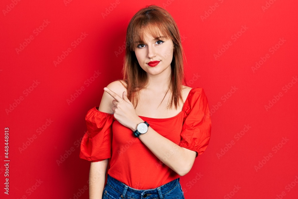 Redhead young woman wearing casual red t shirt pointing with hand finger to the side showing advertisement, serious and calm face Redhead young woman wearing casual red t shirt pointing with hand finger to the side showing advertisement, serious and calm face