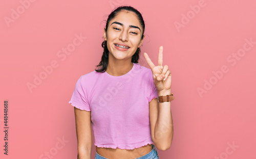Hispanic teenager girl with dental braces wearing casual clothes smiling looking to the camera showing fingers doing victory sign. number two.
