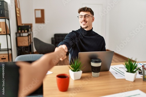 Two business workers smiling happy shaking hands at the office.