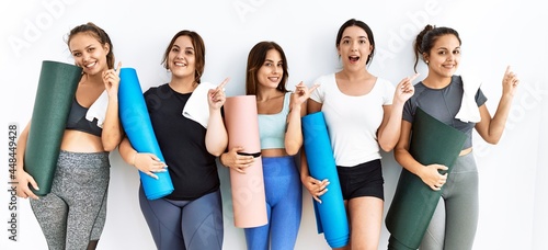 Group of women holding yoga mat standing over isolated background with a big smile on face, pointing with hand finger to the side looking at the camera.