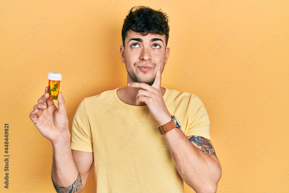 Young hispanic man holding pills serious face thinking about question with hand on chin, thoughtful about confusing idea Young hispanic man holding pills serious face thinking about question with hand on chin, thoughtful about confusing idea