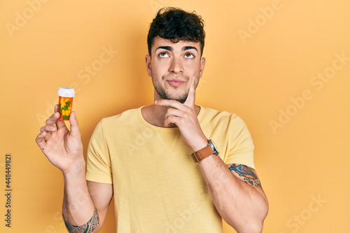 Young hispanic man holding pills serious face thinking about question with hand on chin, thoughtful about confusing idea