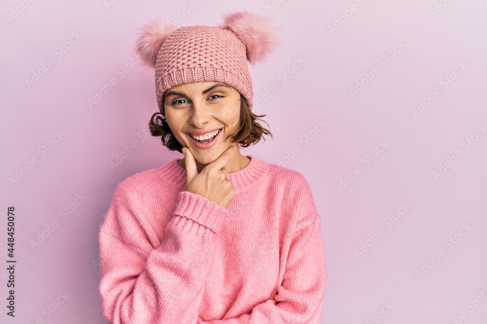 Young brunette woman wearing cute wool cap looking confident at the camera smiling with crossed arms and hand raised on chin. thinking positive. Young brunette woman wearing cute wool cap looking confident at the camera smiling with crossed arms and hand raised on chin. thinking positive.