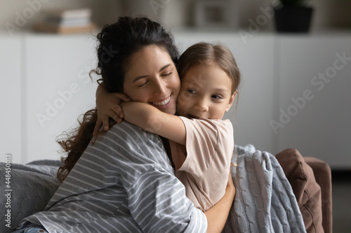 Happy excited mother embracing cute daughter kid with closed eyes. Smiling mom and sweet little girl hugging on couch with love, affection, gratitude. Motherhood, childhood, family