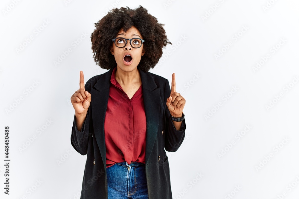 African american woman with afro hair wearing business jacket and glasses amazed and surprised looking up and pointing with fingers and raised arms. African american woman with afro hair wearing business jacket and glasses amazed and surprised looking up and pointing with fingers and raised arms.