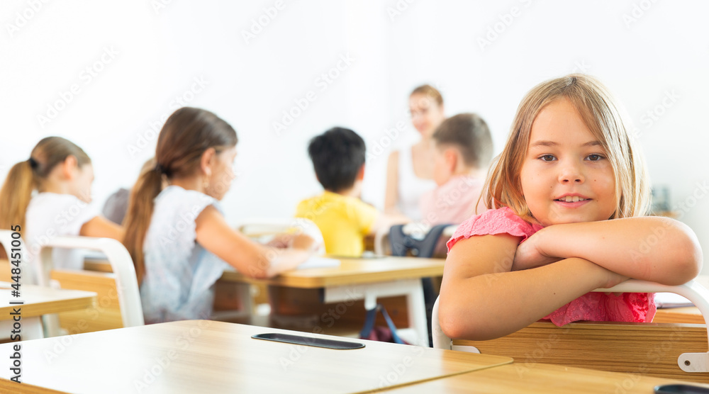 Fototapeta premium Young girl sitting at desk in class room. She have turned around and looking in camera.