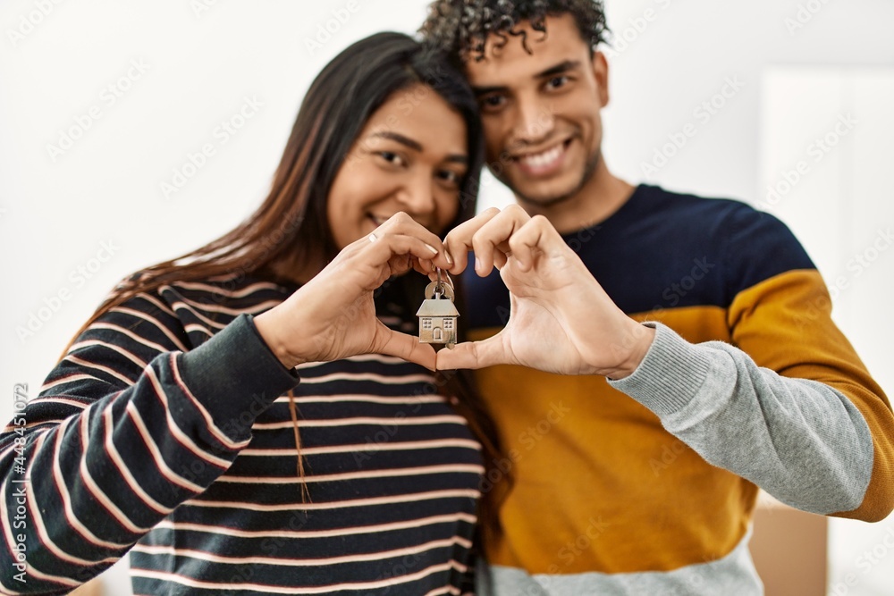 Young latin couple doing heart symbol with hands holding key of new home.