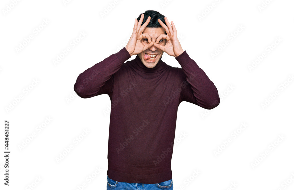 Handsome hispanic man wearing casual turtleneck sweater doing ok gesture like binoculars sticking tongue out, eyes looking through fingers. crazy expression. Handsome hispanic man wearing casual turtleneck sweater doing ok gesture like binoculars sticking tongue out, eyes looking through fingers. crazy expression.