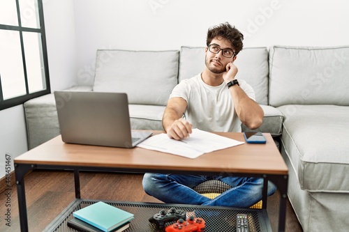 Hispanic man doing papers at home with hand on chin thinking about question, pensive expression. smiling with thoughtful face. doubt concept.