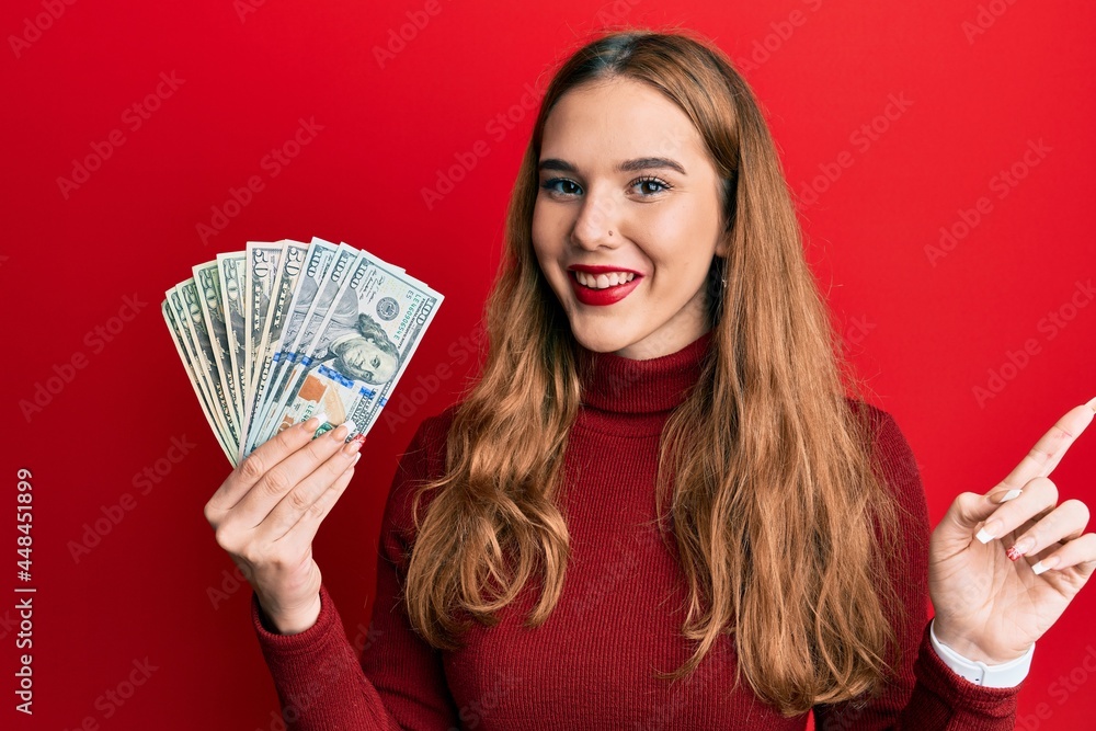 Young blonde woman holding dollars smiling happy pointing with hand and finger to the side Young blonde woman holding dollars smiling happy pointing with hand and finger to the side