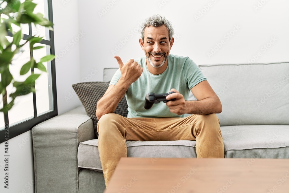 Middle age hispanic man playing video game sitting on the sofa pointing thumb up to the side smiling happy with open mouth Middle age hispanic man playing video game sitting on the sofa pointing thumb up to the side smiling happy with open mouth