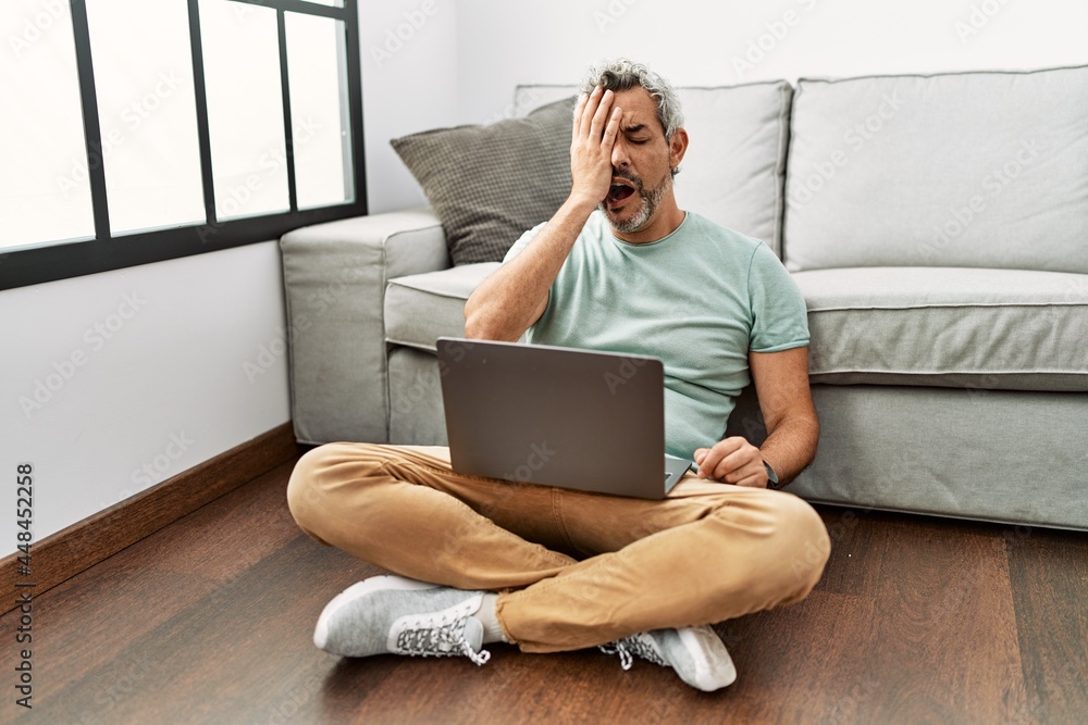 Middle age hispanic man using laptop sitting on the floor at the living room yawning tired covering half face, eye and mouth with hand. face hurts in pain. Middle age hispanic man using laptop sitting on the floor at the living room yawning tired covering half face, eye and mouth with hand. face hurts in pain.