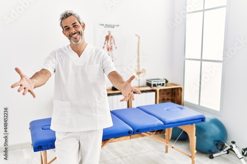 Middle age hispanic therapist man working at pain recovery clinic looking at the camera smiling with open arms for hug. cheerful expression embracing happiness.