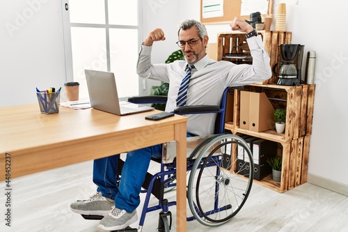 Middle age hispanic man working at the office sitting on wheelchair showing arms muscles smiling proud. fitness concept.