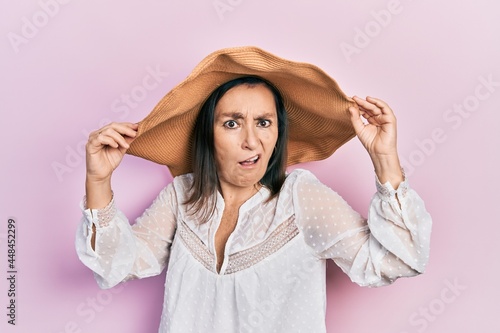 Middle age hispanic woman wearing summer hat in shock face, looking skeptical and sarcastic, surprised with open mouth