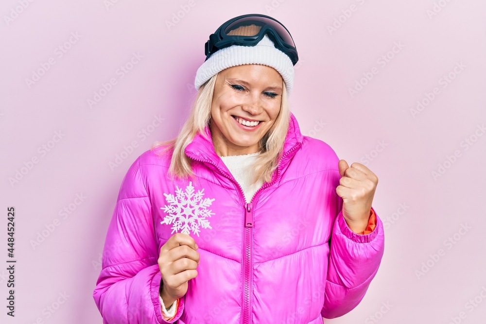 Beautiful caucasian blonde woman wearing snow wear and sky glasses holding snowflake screaming proud, celebrating victory and success very excited with raised arm Beautiful caucasian blonde woman wearing snow wear and sky glasses holding snowflake screaming proud, celebrating victory and success very excited with raised arm