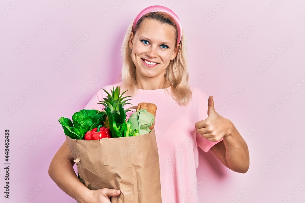 Beautiful caucasian blonde woman holding paper bag with groceries smiling happy and positive, thumb up doing excellent and approval sign Beautiful caucasian blonde woman holding paper bag with groceries smiling happy and positive, thumb up doing excellent and approval sign