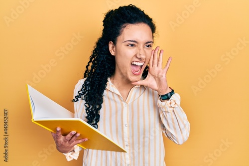 Young hispanic woman with curly hair holding book shouting and screaming loud to side with hand on mouth. communication concept.