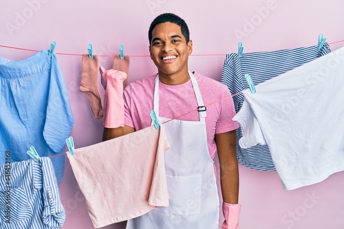 Young handsome hispanic man wearing cleaner apron holding clothes on clothesline smiling with happy face looking and pointing to the side with thumb up.