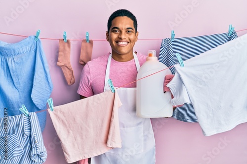 Young handsome hispanic man doing laundry holding detergent bottle looking positive and happy standing and smiling with a confident smile showing teeth