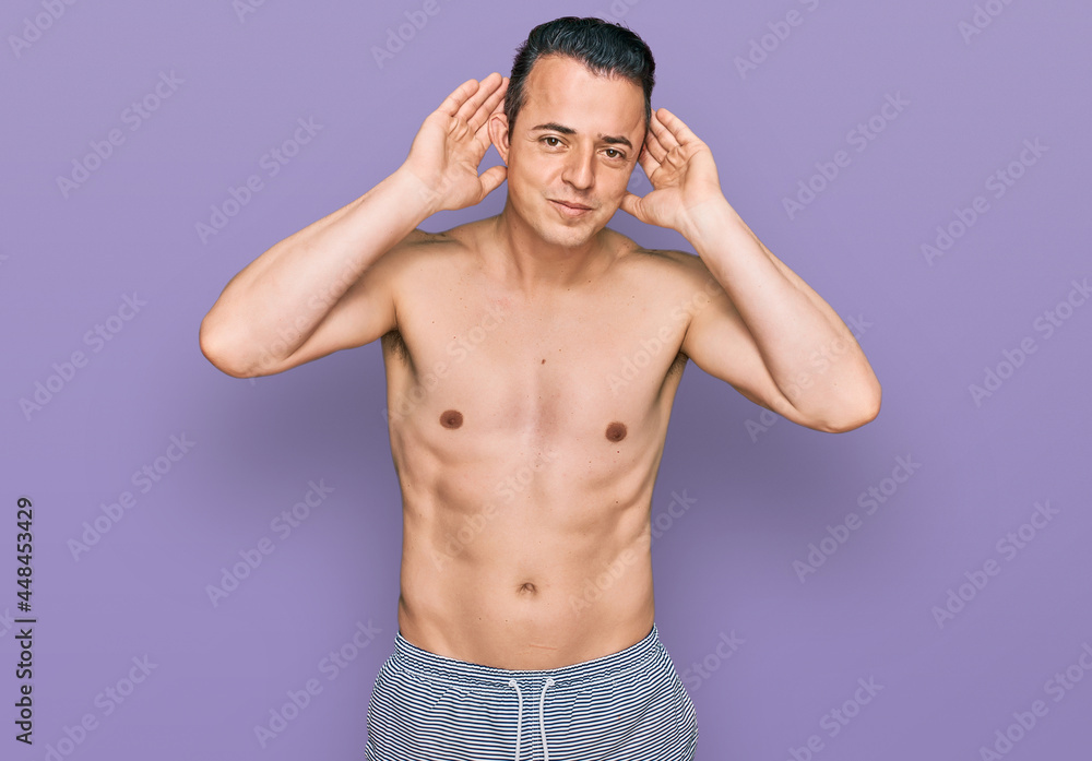 Fototapeta premium Handsome young man wearing swimwear shirtless trying to hear both hands on ear gesture, curious for gossip. hearing problem, deaf
