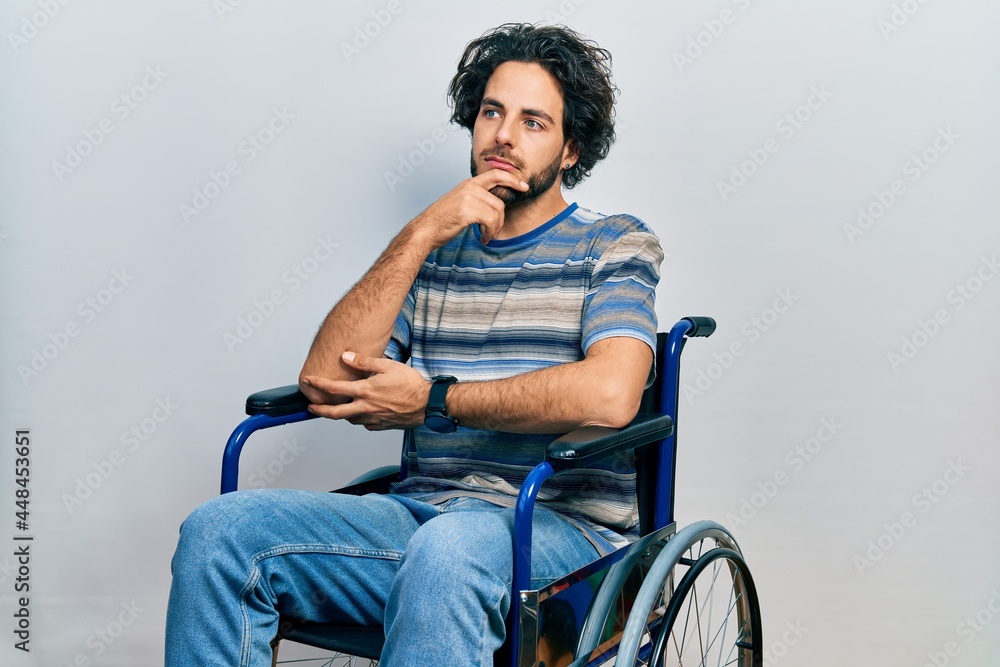 Handsome hispanic man sitting on wheelchair with hand on chin thinking about question, pensive expression. smiling with thoughtful face. doubt concept. Handsome hispanic man sitting on wheelchair with hand on chin thinking about question, pensive expression. smiling with thoughtful face. doubt concept.