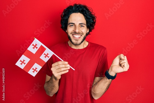 Handsome hispanic man holding georgia flag screaming proud, celebrating victory and success very excited with raised arm