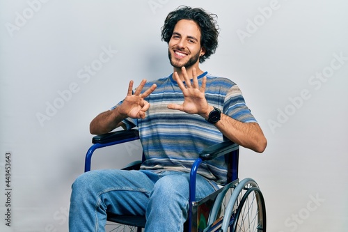 Handsome hispanic man sitting on wheelchair showing and pointing up with fingers number eight while smiling confident and happy.