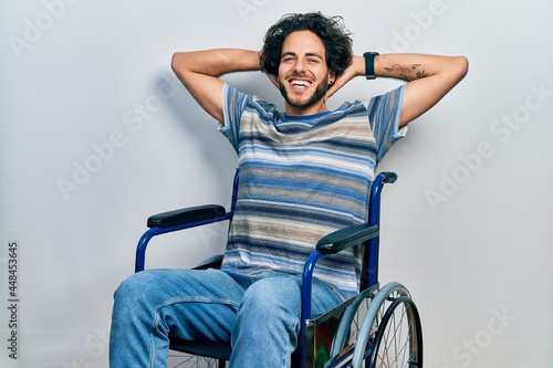 Handsome hispanic man sitting on wheelchair relaxing and stretching, arms and hands behind head and neck smiling happy