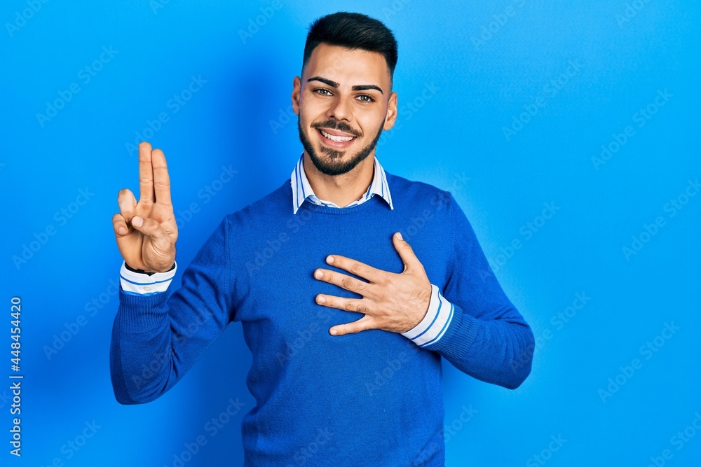 Young hispanic man with beard wearing casual blue sweater smiling swearing with hand on chest and fingers up, making a loyalty promise oath Young hispanic man with beard wearing casual blue sweater smiling swearing with hand on chest and fingers up, making a loyalty promise oath